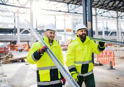Twee technici in fluogele werkkleding dragen samen een metalen profielbalk door een industriële hal